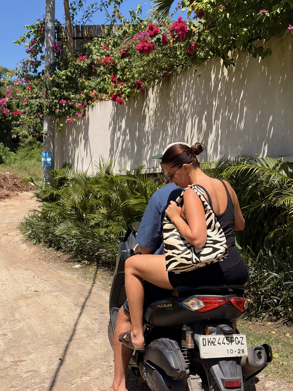 Two people riding a scooter on a dirt path with flowers and greenery around. Woman holding a zebra cowhide shoulder bag.