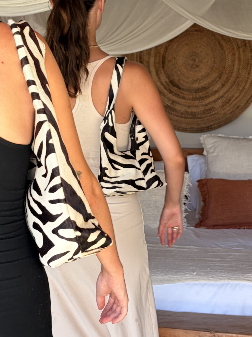 Two women wearing neutral dressed with zebra pattern cowhide shoulderbag in a bedroom setting.